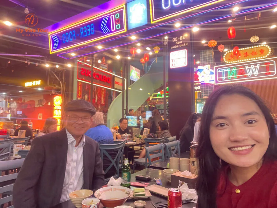 Tito and Rachel at a lively neon-lit street-food spot in Hanoi's Old Quarter at night
