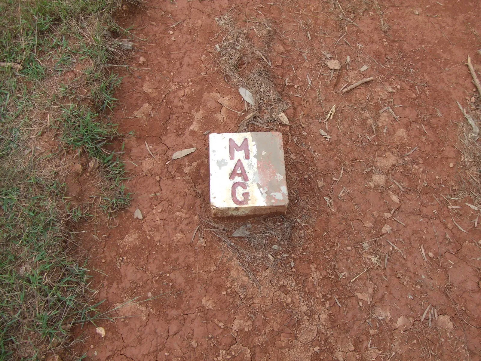 A white MAG concrete marker on red earth showing ground cleared of unexploded ordnance at a Plain of Jars site near Phonsavan