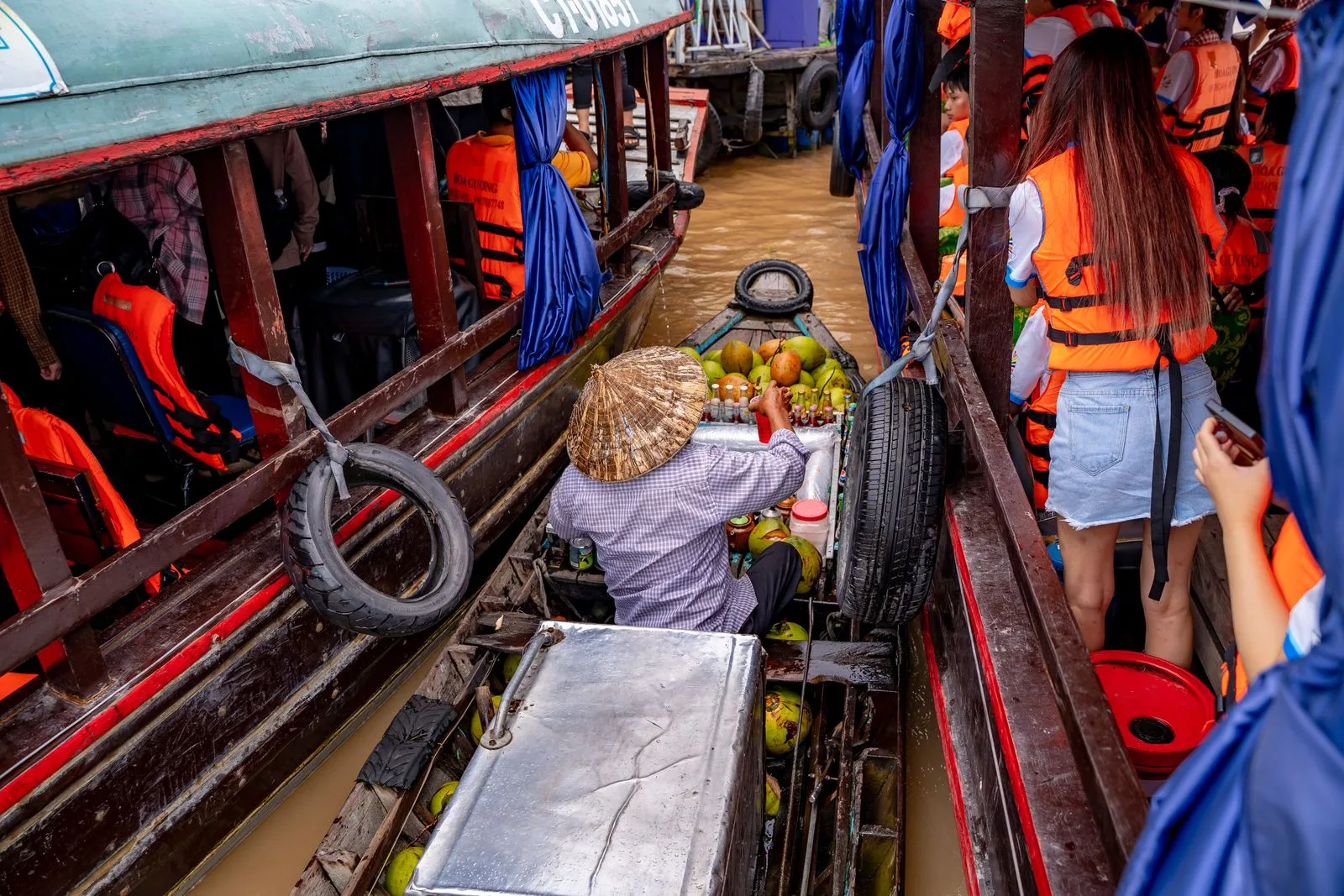 A vendor in a conical hat selling coconuts and drinks between tour boats at Cai Rang floating market near Can Tho