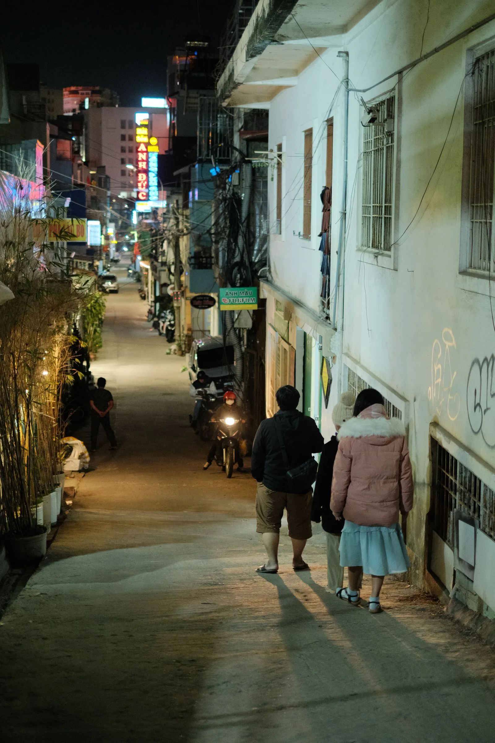 People in warm jackets walking a Da Lat street at night under glowing neon shop signs