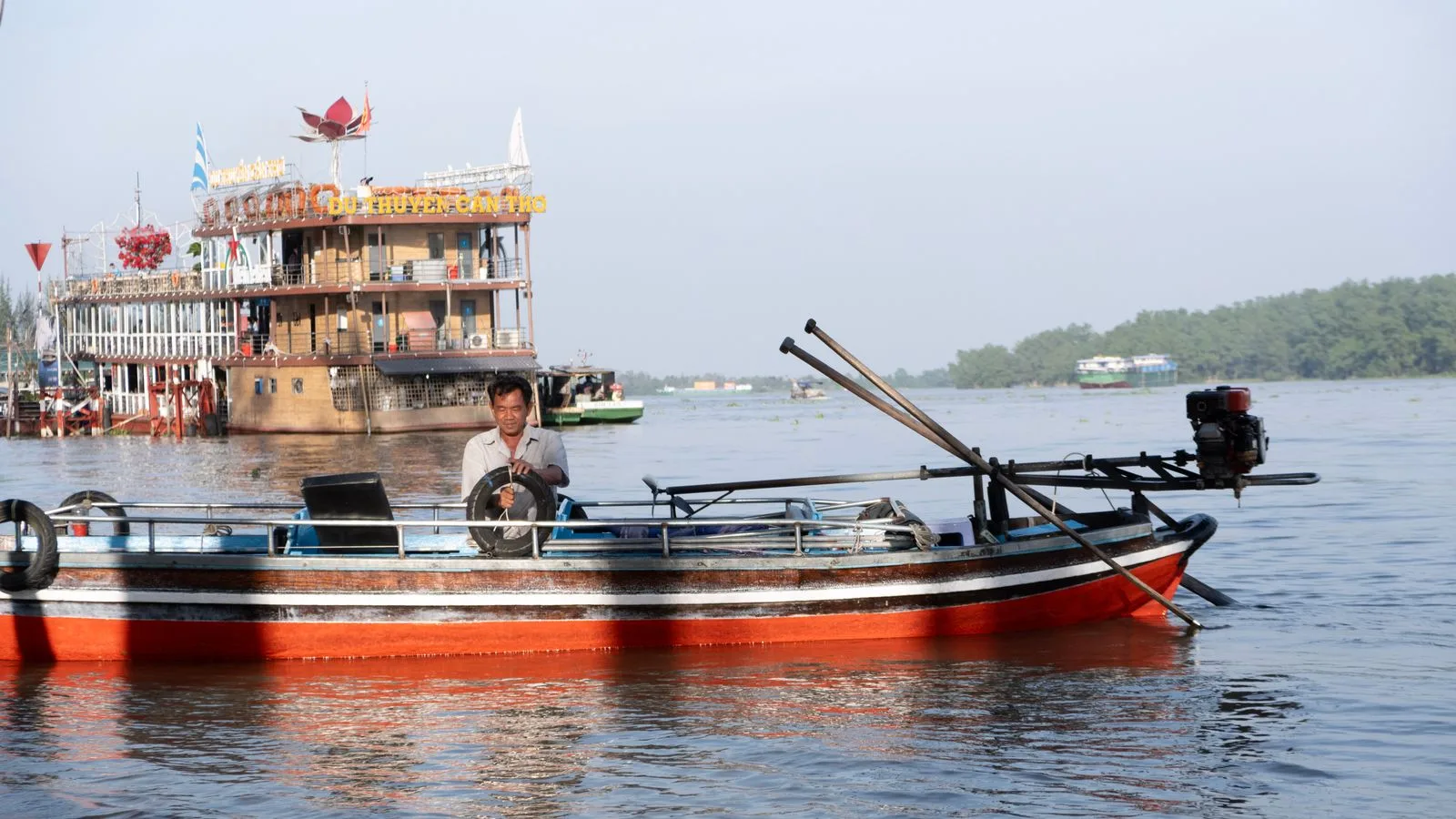 A local boatman steering a red wooden boat on the Can Tho river with a river cruise boat behind