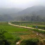 Pu Luong travel guide: trekkers on a path through bright green rice paddies below misty karst hills, with a thatched stilt house in a Pu Luong Nature Reserve valley