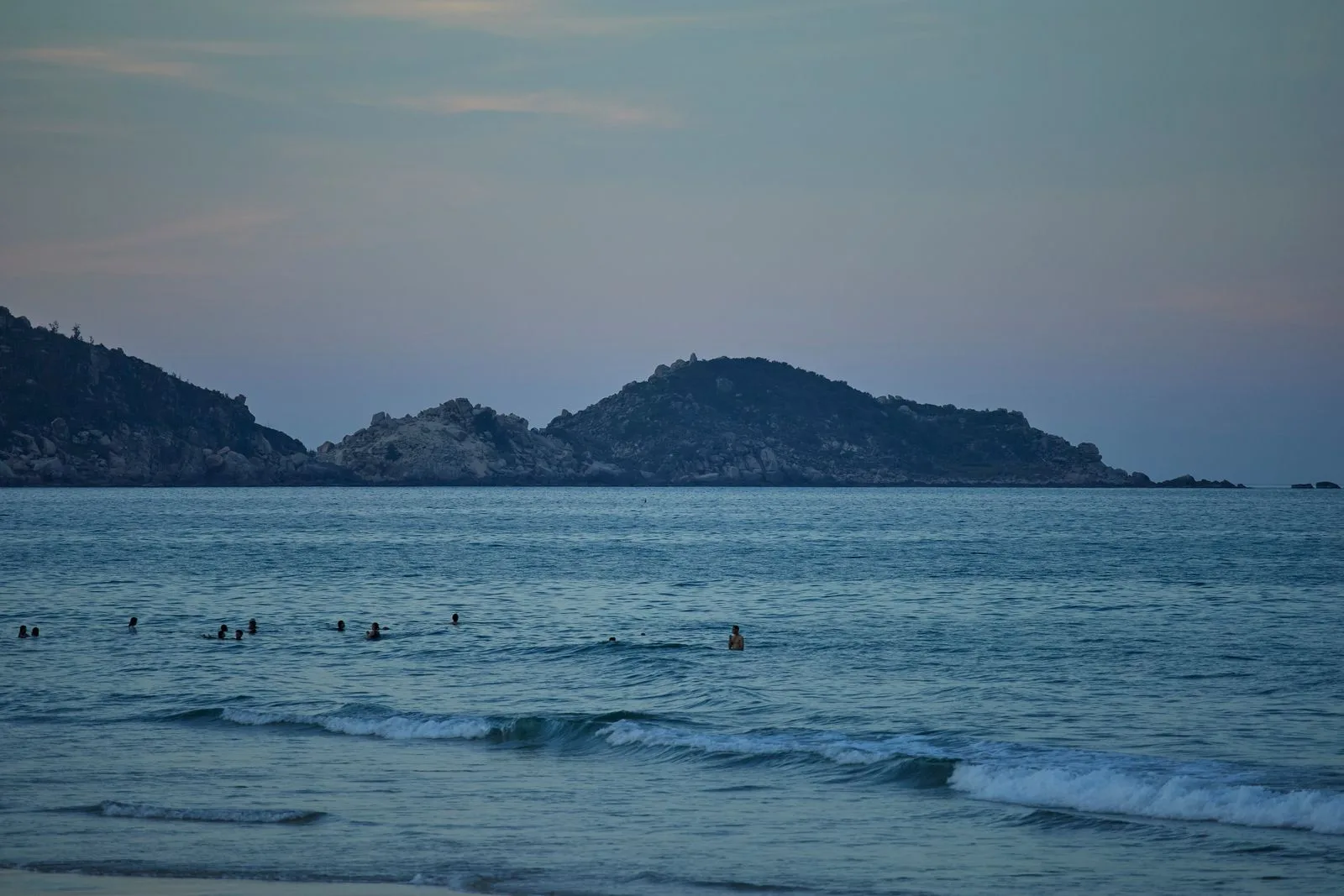 Swimmers in calm sea at dusk off a Quy Nhon beach with a rocky island on the horizon