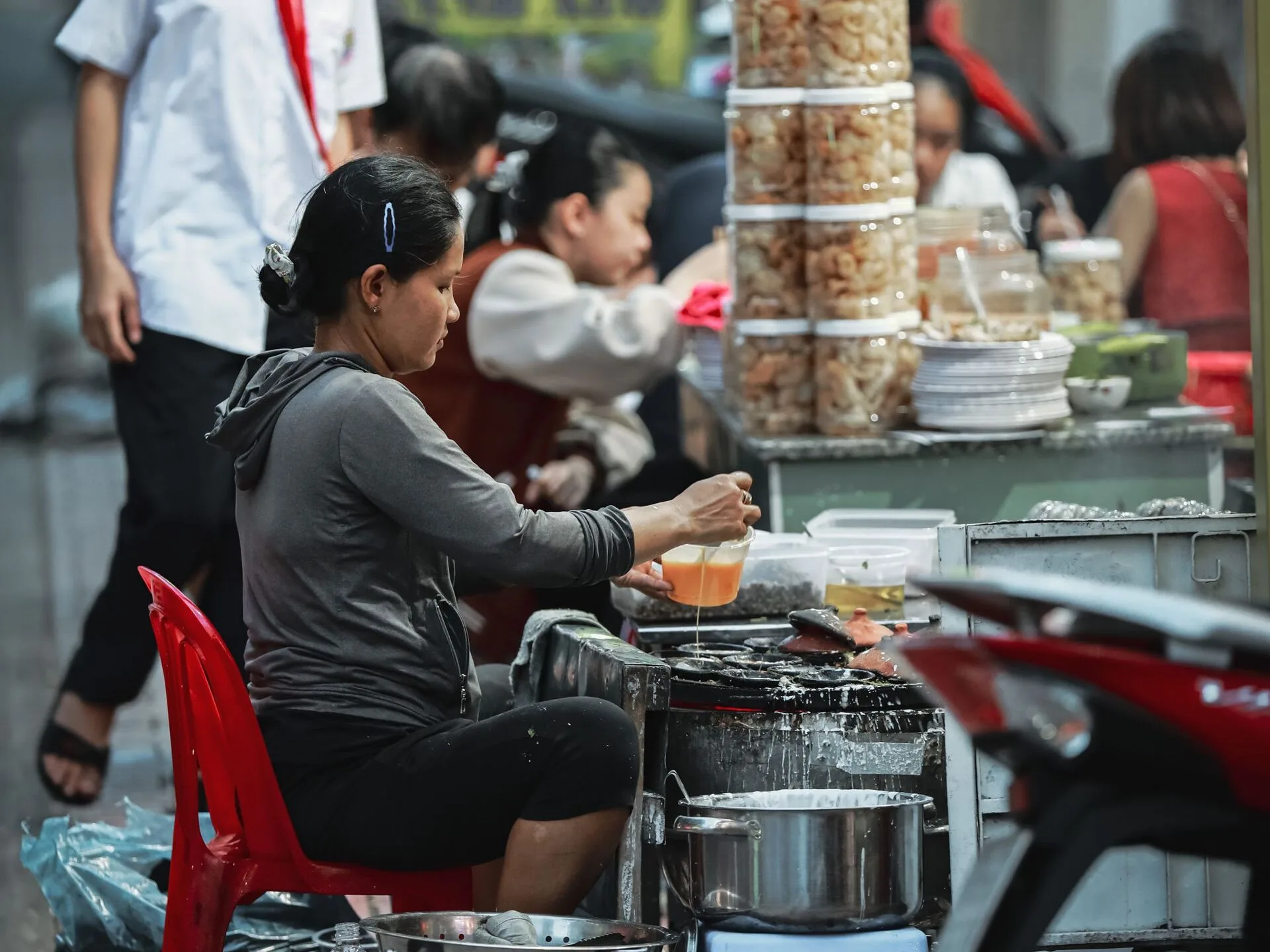 Street vendor in Nha Trang pouring batter into a pan of round moulds to cook banh can, beside jars of crackers and stacked plates