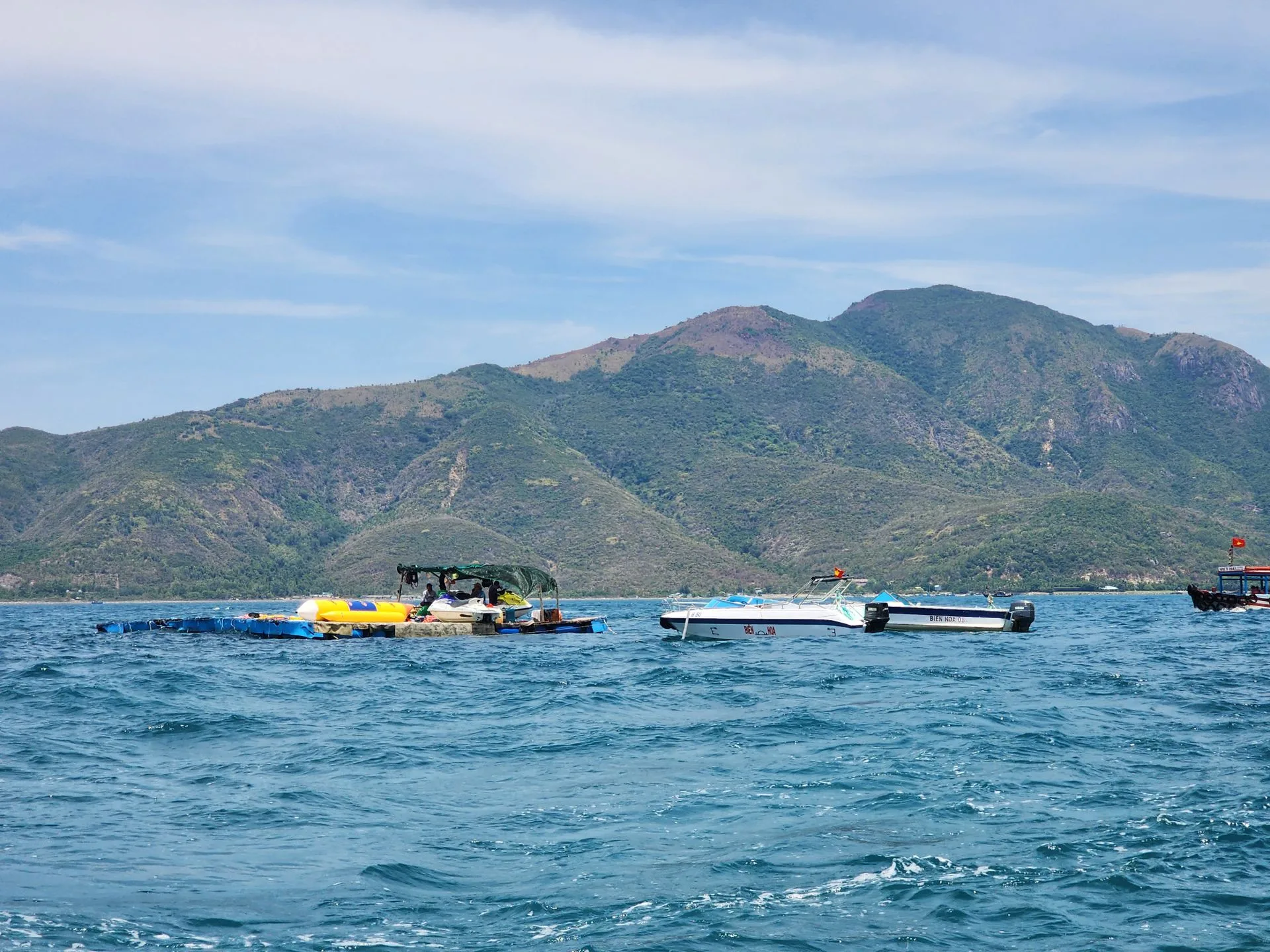 Speedboats, jet skis and a banana boat moored on a floating platform in Nha Trang Bay with a green mountain headland behind