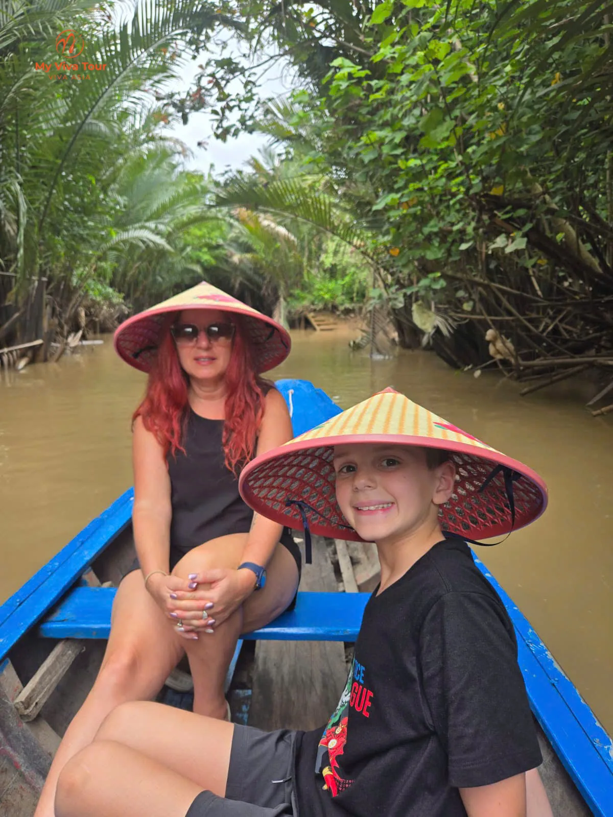 Mother and son in conical hats on a wooden sampan gliding along a palm-shaded waterway in Vietnam