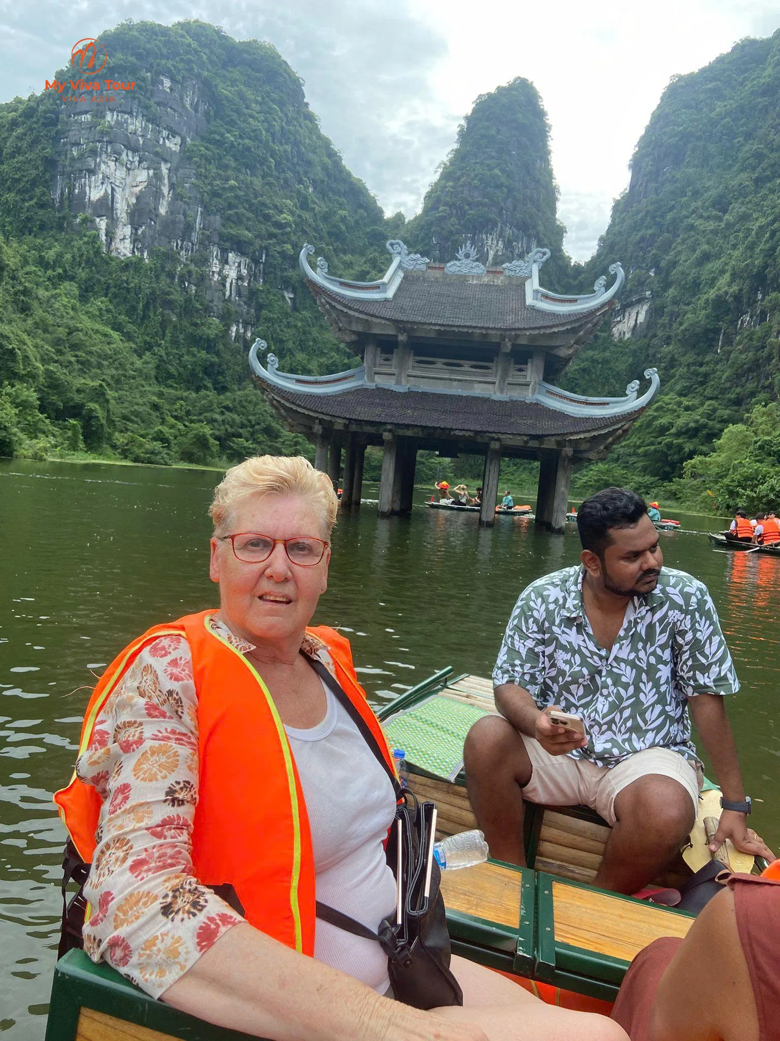 Missy in a life jacket on a Trang An rowing boat, a two-tier pavilion and limestone karsts behind, Ninh Binh