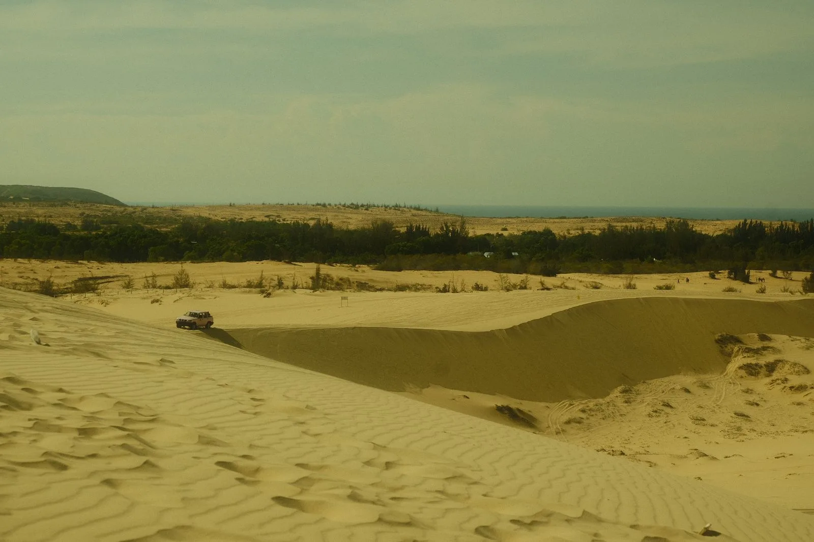 Rippled pale sand dunes near Mui Ne with a jeep crossing them and the sea on the horizon