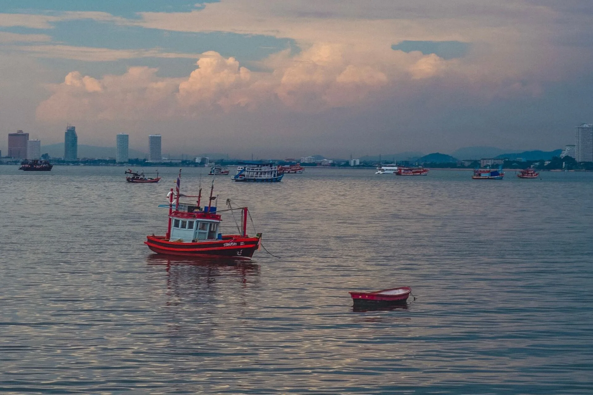 Red wooden fishing boat moored in Pattaya Bay at dusk with the city skyline on the horizon